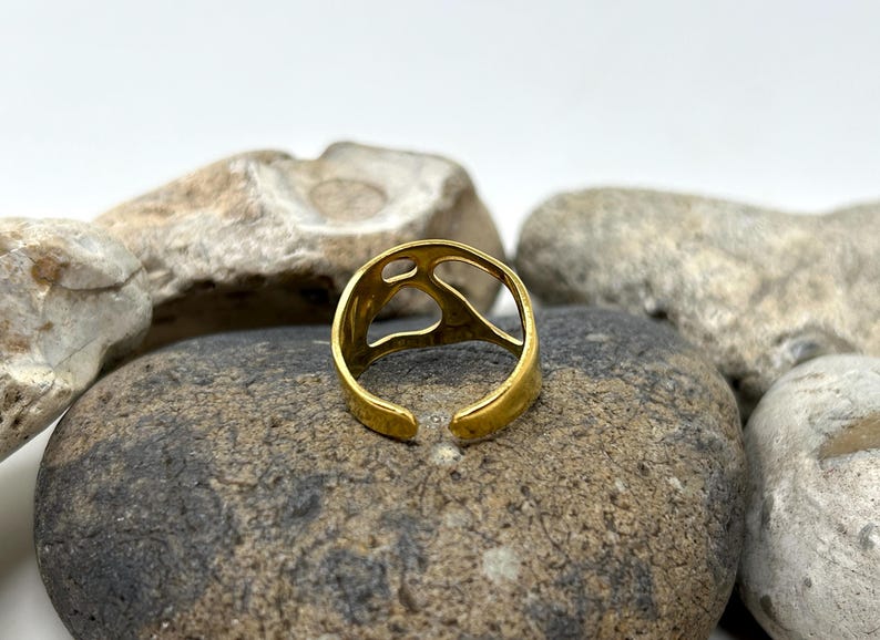 May include: A gold-colored adjustable ring with an open design. The ring is set against a backdrop of gray and tan stones. The ring's design features an open, abstract shape.