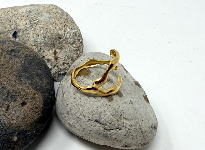 Puede incluir: Un anillo dorado de forma abstracta descansa sobre una piedra gris. El anillo tiene un dise&ntilde;o org&aacute;nico y fluido con l&iacute;neas curvas y una superficie texturizada. El fondo presenta dos piedras grises adicionales y una superficie blanca.