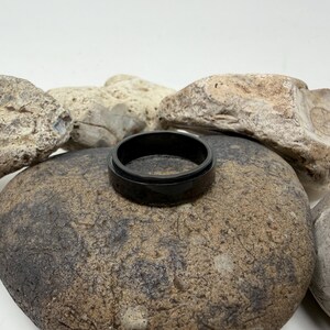 May include: A black ring with a glossy finish rests on a large, textured stone. The ring's dark color contrasts with the neutral tones of the surrounding rocks, creating a simple yet striking composition. The ring appears to be made of a solid material.