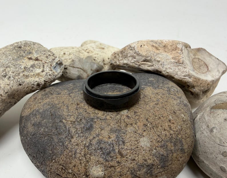 May include: A black ring with a glossy finish rests on a large, textured stone. The ring's dark color contrasts with the neutral tones of the surrounding rocks, creating a simple yet striking composition. The ring appears to be made of a solid material.