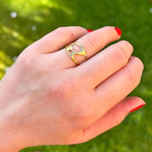 May include: A gold-colored ring with an open, abstract design is worn on a finger. The ring's shape features irregular cutouts. The hand is positioned against a blurred green background, with red nail polish on the fingernails.