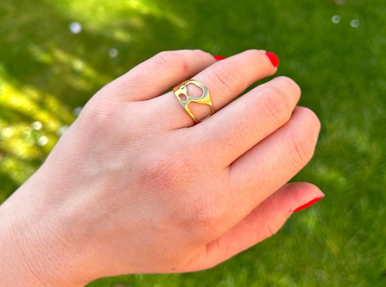 May include: A gold-colored ring with an open, abstract design is worn on a finger. The ring's shape features irregular cutouts. The hand is positioned against a blurred green background, with red nail polish on the fingernails.