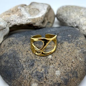 May include: A gold-colored ring with an open, abstract design. The ring is set against a backdrop of gray and tan stones. The ring's design features a series of interconnected, organic shapes, creating an open and airy aesthetic. The ring is a piece of jewelry.