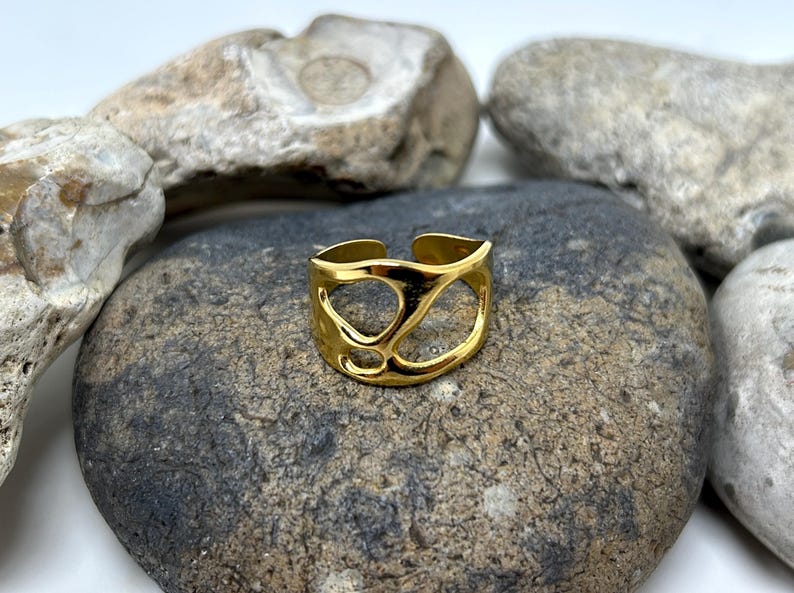 May include: A gold-colored ring with an open, abstract design. The ring is set against a backdrop of gray and tan stones. The ring's design features a series of interconnected, organic shapes, creating an open and airy aesthetic. The ring is a piece of jewelry.