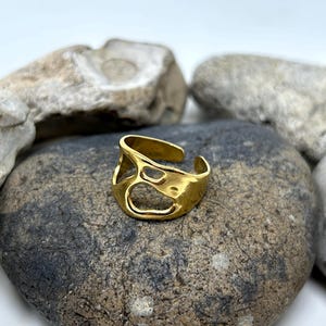 May include: A gold-colored adjustable ring with an open, organic design. The ring features irregular cutouts and sits on a gray stone, with other stones in the background. The ring is a piece of jewelry.