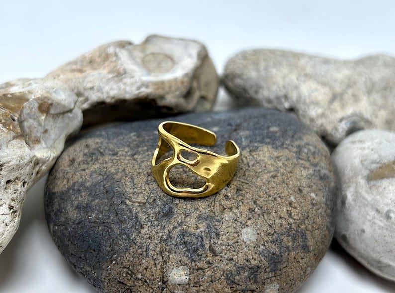 May include: A gold-colored adjustable ring with an open, organic design. The ring features irregular cutouts and sits on a gray stone, with other stones in the background. The ring is a piece of jewelry.