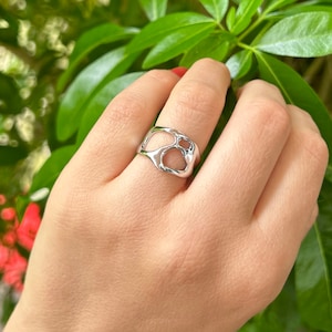May include: A silver ring with an organic, openwork design. The ring features a series of interconnected, irregular shapes, creating a unique and modern aesthetic. The ring is shown on a hand against a backdrop of green foliage.