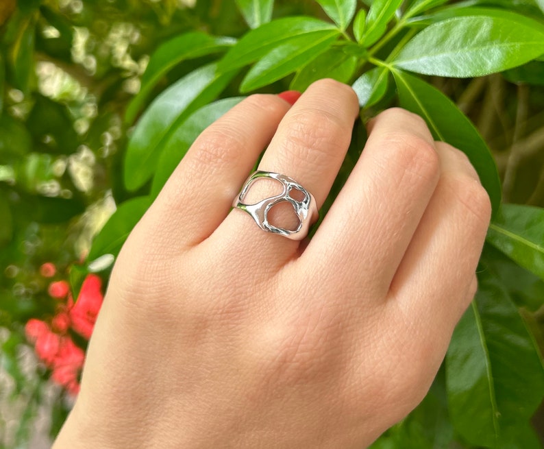 May include: A silver ring with an organic, openwork design. The ring features a series of interconnected, irregular shapes, creating a unique and modern aesthetic. The ring is shown on a hand against a backdrop of green foliage.