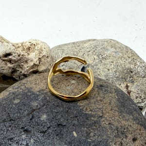 Puede incluir: Un anillo dorado con un dise&ntilde;o org&aacute;nico y fluido. El anillo se presenta sobre un fondo de piedras grises y beige, creando un contraste natural y texturizado. El dise&ntilde;o abierto del anillo a&ntilde;ade un toque moderno a la joya.