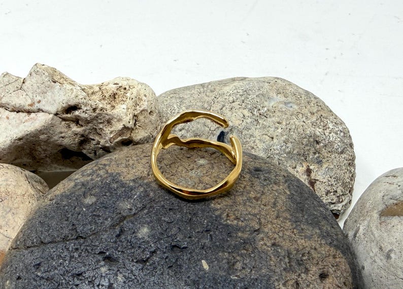 Puede incluir: Un anillo dorado con un dise&ntilde;o org&aacute;nico y fluido. El anillo se presenta sobre un fondo de piedras grises y beige, creando un contraste natural y texturizado. El dise&ntilde;o abierto del anillo a&ntilde;ade un toque moderno a la joya.