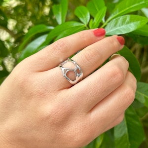 May include: A silver-colored ring with an open, organic design. The ring features irregular, interconnected shapes, creating a unique and modern aesthetic. The ring is shown on a hand with red nail polish, set against a backdrop of green leaves.