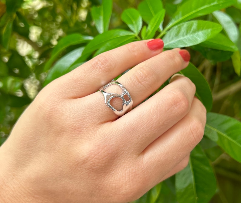 May include: A silver-colored ring with an open, organic design. The ring features irregular, interconnected shapes, creating a unique and modern aesthetic. The ring is shown on a hand with red nail polish, set against a backdrop of green leaves.