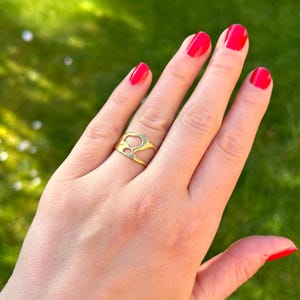 May include: A gold-colored ring with an abstract design is worn on a hand with red nail polish. The ring has an open, irregular shape. The background is a blurred green lawn.