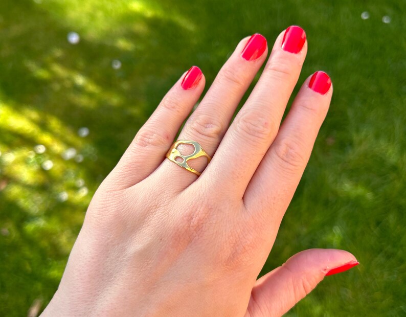 May include: A gold-colored ring with an abstract design is worn on a hand with red nail polish. The ring has an open, irregular shape. The background is a blurred green lawn.