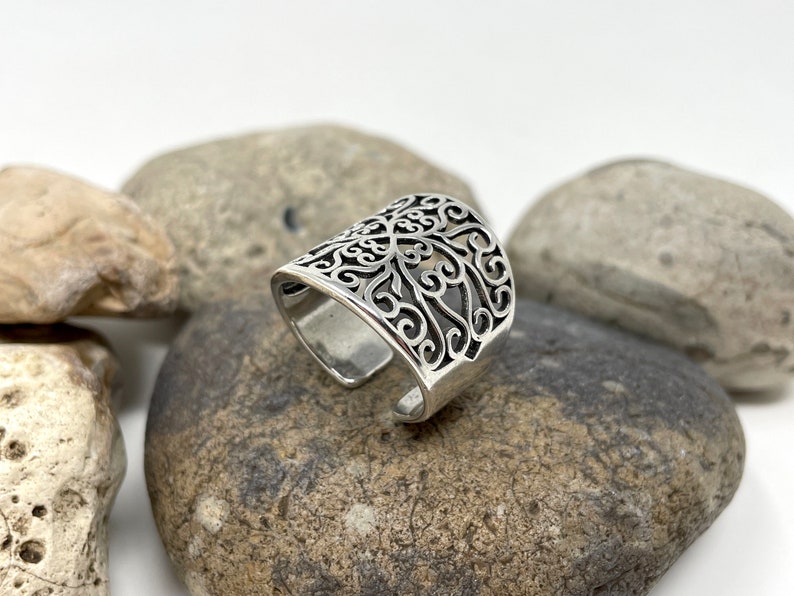 May include: A silver ring with a filigree design. The ring is open and adjustable.
