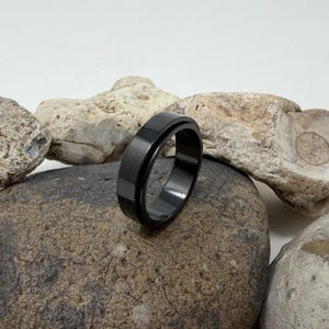 May include: A black ring with a flat, matte finish. The ring is set against a backdrop of various rocks, showcasing its sleek design. The ring's dark color contrasts with the lighter tones of the stones.