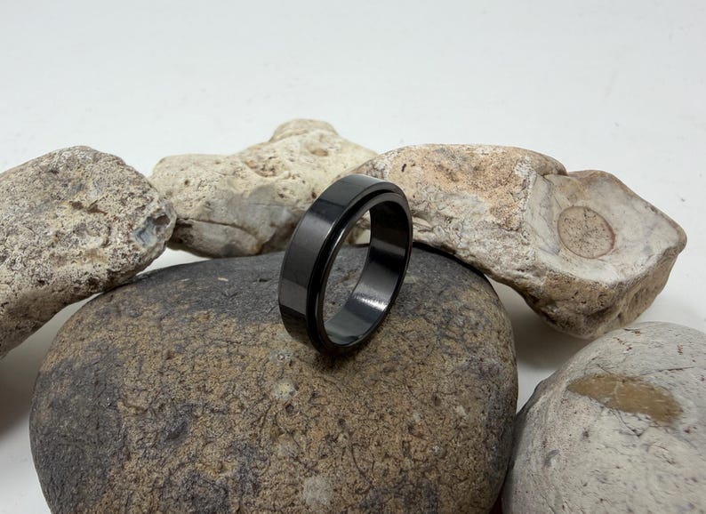 May include: A black ring with a flat, matte finish. The ring is set against a backdrop of various rocks, showcasing its sleek design. The ring's dark color contrasts with the lighter tones of the stones.