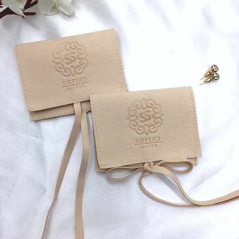 May include: Two beige suede jewelry pouches with the text "SOFIA'S SILVER" and a decorative logo. The pouches are tied with matching suede ribbons. A pair of gold stud earrings is visible in the background.