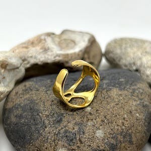 May include: A gold-colored open ring with an abstract design. The ring is set on a gray stone, with other stones in the background. The ring has a smooth, polished finish and a unique, modern style.