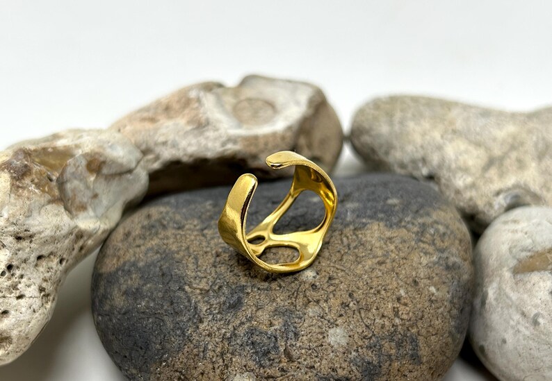 May include: A gold-colored open ring with an abstract design. The ring is set on a gray stone, with other stones in the background. The ring has a smooth, polished finish and a unique, modern style.