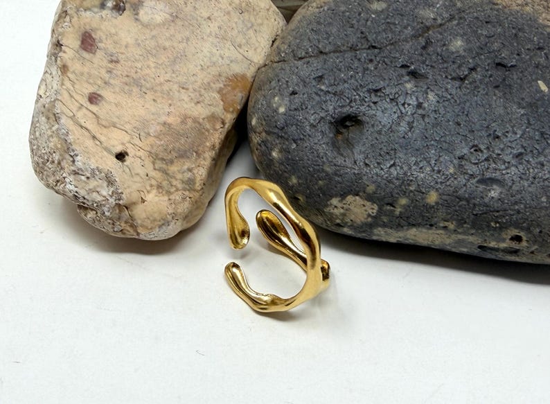 Puede incluir: Un anillo dorado con un dise&ntilde;o org&aacute;nico y fluido. El anillo tiene una forma abierta &uacute;nica con l&iacute;neas suaves y curvas. El anillo se muestra sobre una superficie blanca, con dos piedras en el fondo.