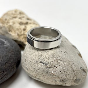 May include: A silver-colored spinner ring with a black accent band. The ring is displayed on a light gray stone, with other stones in the background. The ring is a simple, modern design.