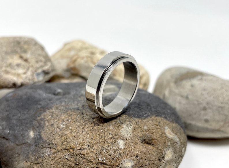 May include: A silver-colored ring with a flat, wide band and a grooved outer edge. The ring is made of a shiny, metallic material and is displayed on a background of gray and brown stones. The ring's design is simple and modern.