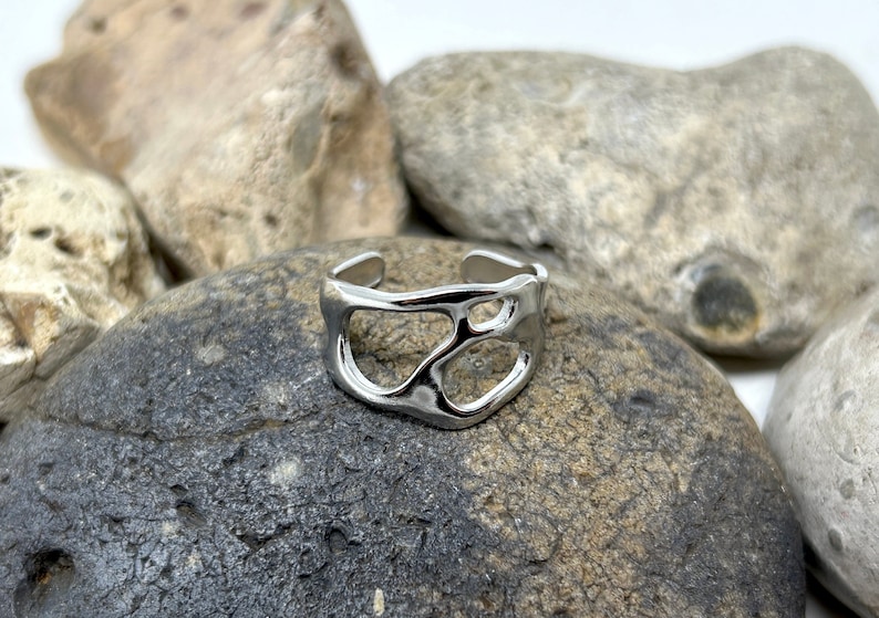 May include: A silver-colored, open-design ring with an irregular, organic shape. The ring is set against a backdrop of gray and tan rocks, creating a natural contrast. The ring's adjustable design allows for a comfortable fit.