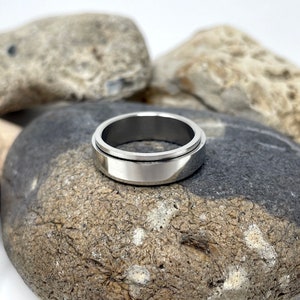 May include: A silver-colored, stainless steel spinner ring. The ring has a wide band with a rotating outer ring. The ring is displayed on a gray and black stone, with other stones in the background. The ring is a piece of jewelry.