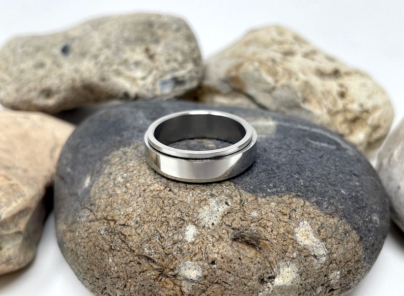 May include: A silver-colored, stainless steel spinner ring. The ring has a wide band with a rotating outer ring. The ring is displayed on a gray and black stone, with other stones in the background. The ring is a piece of jewelry.