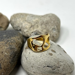 May include: A gold-colored ring with an abstract design featuring irregular cutouts. The ring is displayed on a gray stone, with other stones in the background. The ring's design is open and airy.