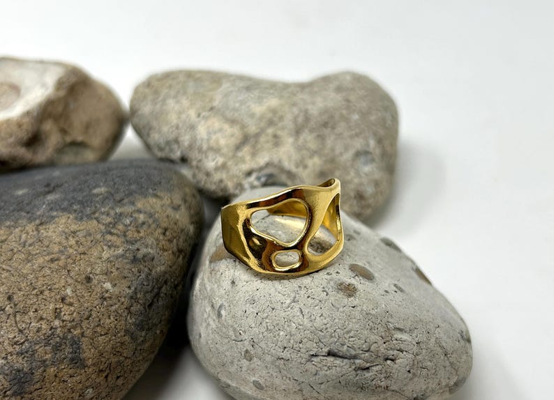 May include: A gold-colored ring with an abstract design featuring irregular cutouts. The ring is displayed on a gray stone, with other stones in the background. The ring's design is open and airy.