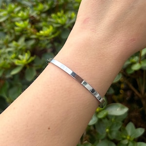 May include: A silver bangle bracelet worn on a person's wrist. The bracelet is simple and sleek, with a smooth, polished finish.