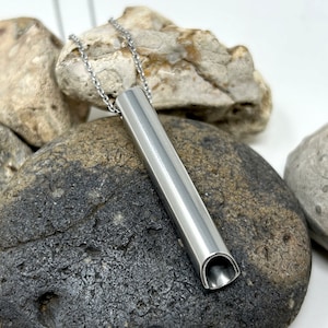 May include: A silver pendant necklace with a hollow, cylindrical charm. The charm is smooth and has a rounded opening at the bottom. The necklace is displayed on a bed of gray rocks.
