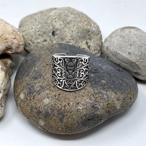 May include: A silver ring with a filigree design. The ring is on a gray rock.