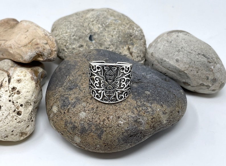 May include: A silver ring with a filigree design. The ring is on a gray rock.