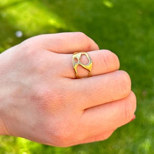 May include: A gold-colored ring with an abstract design. The ring features open spaces and a smooth, polished surface. The ring is worn on a finger, with a green background visible.