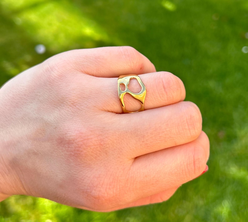 May include: A gold-colored ring with an abstract design. The ring features open spaces and a smooth, polished surface. The ring is worn on a finger, with a green background visible.
