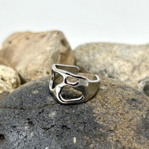 May include: A silver-colored ring with an open, abstract design. The ring features irregular cutouts and a smooth, polished surface. The adjustable band allows for a comfortable fit. The ring is displayed on a dark gray rock.