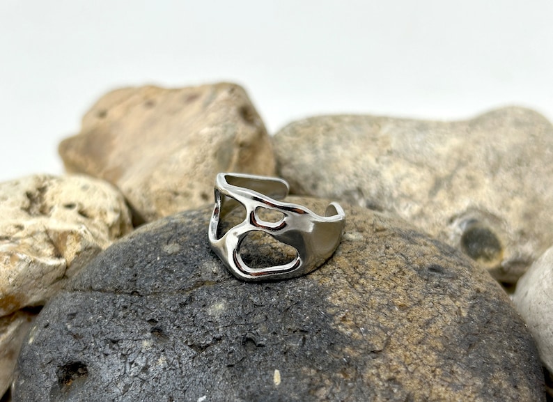 May include: A silver-colored ring with an open, abstract design. The ring features irregular cutouts and a smooth, polished surface. The adjustable band allows for a comfortable fit. The ring is displayed on a dark gray rock.