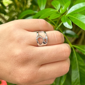 May include: A silver-colored ring with an open, organic design. The ring features irregular cut-outs, creating a unique and modern aesthetic. The ring is shown on a hand, with a blurred green background.