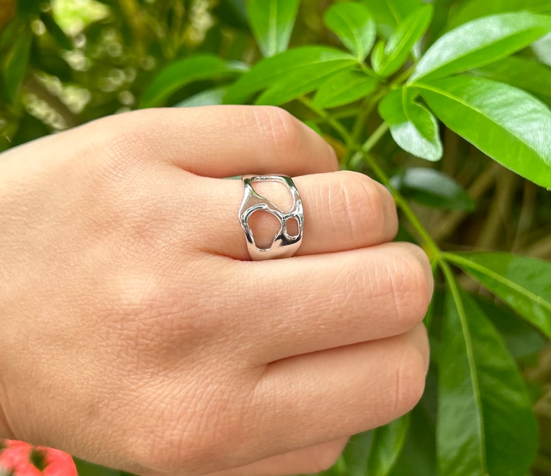 May include: A silver-colored ring with an open, organic design. The ring features irregular cut-outs, creating a unique and modern aesthetic. The ring is shown on a hand, with a blurred green background.