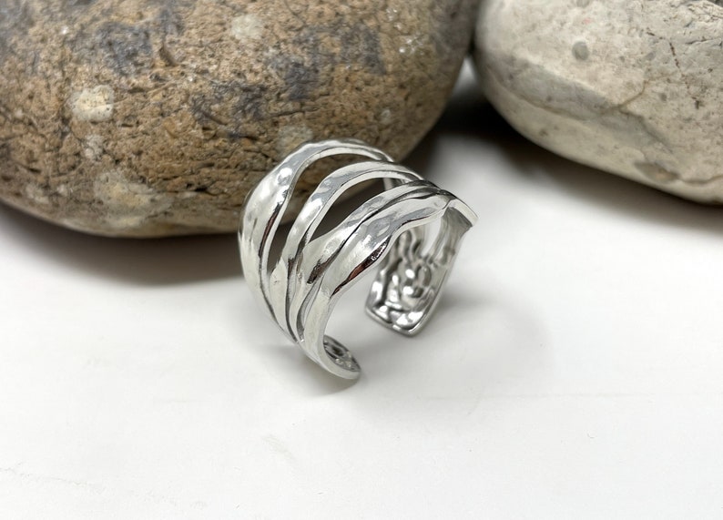 May include: A silver ring with a wavy, textured design. The ring is open and adjustable.