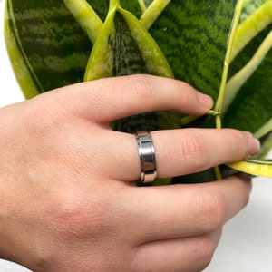May include: A silver-colored ring with a grooved design. The ring is held in a hand near a green and yellow plant. The ring has a flat top and a polished finish, with a band of grooves around the center.