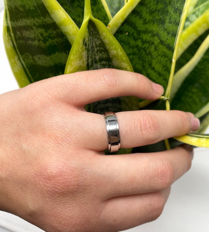 May include: A silver-colored ring with a grooved design. The ring is held in a hand near a green and yellow plant. The ring has a flat top and a polished finish, with a band of grooves around the center.