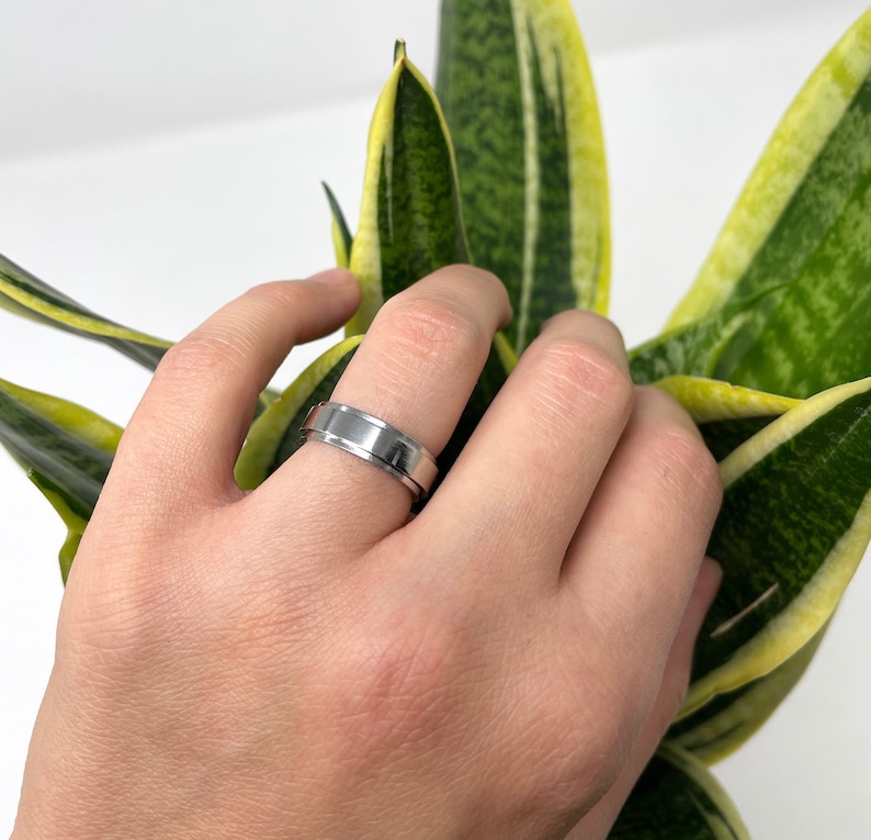 May include: A silver-colored ring with a grooved center band. The ring is on a finger, with a plant in the background. The ring has a polished finish and a simple, modern design.