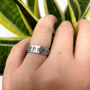 May include: A silver ring with a repeating pattern of small, black flower designs around the band.