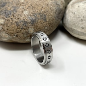 May include: A silver spinning ring with a black flower design. The ring is on a white surface with two rocks behind it.