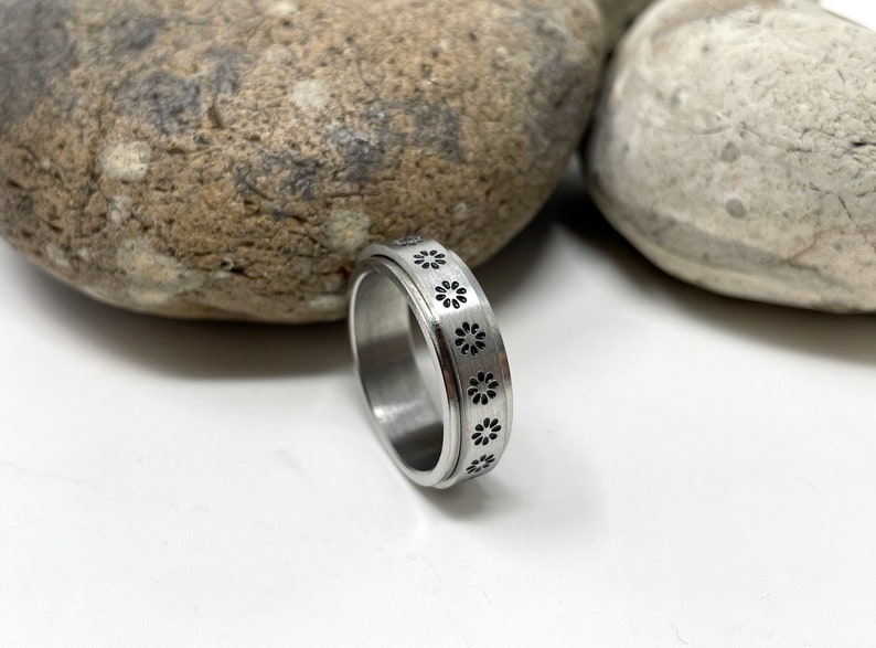 May include: A silver spinning ring with a black flower design. The ring is on a white surface with two rocks behind it.