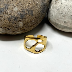 May include: A gold-colored adjustable ring with an open, abstract design. The ring features irregular cutouts, creating an artistic and modern look. The ring is displayed on a white surface, with two large, textured stones in the background.